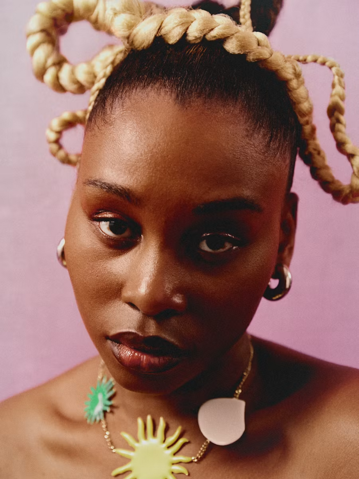 Black woman with sculptural blonde looped braids and gold hoop earrings against a pink background